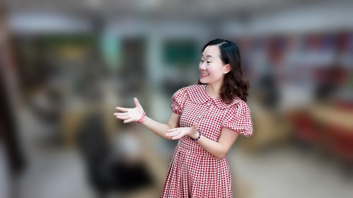 Thảo váy caro đỏ đưa hai tay như giới thiệu, mỉm cười tại văn phòng Tâm - Tầm - Tài tại văn phòng Tâm - Tầm - Tài