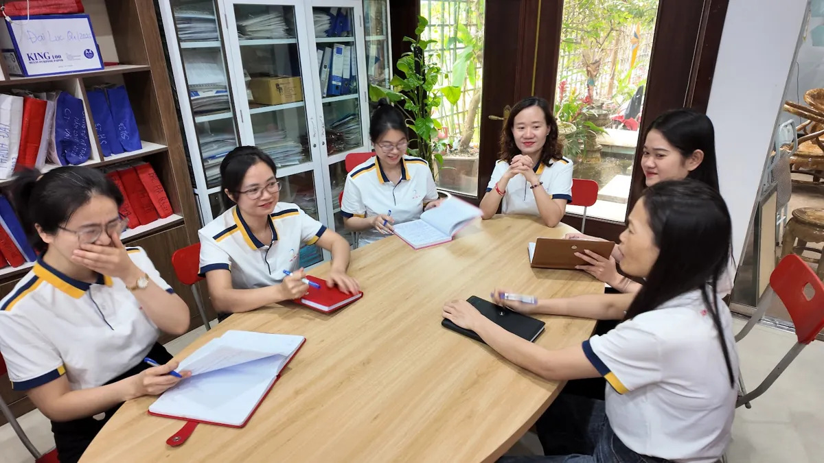 Nhân viên đồng phục họp quanh bàn dài, ghi chép sổ tay