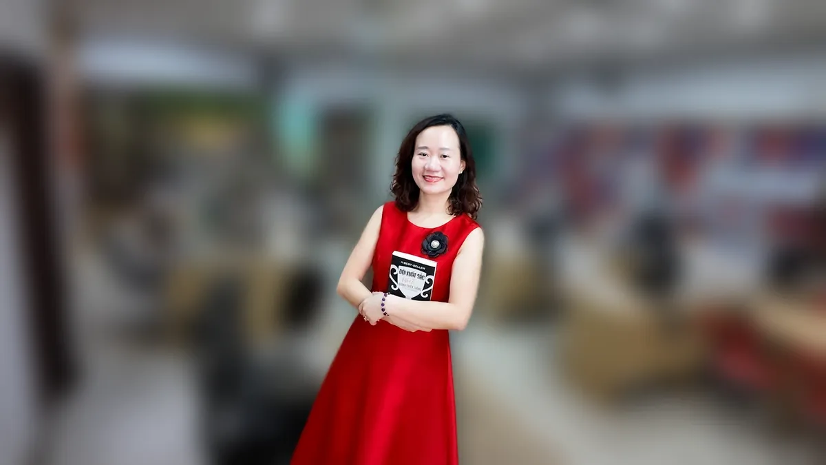 Thảo váy caro đỏ đưa hai tay như giới thiệu, mỉm cười tại văn phòng Tâm - Tầm - Tài tại văn phòng Tâm - Tầm - Tài
